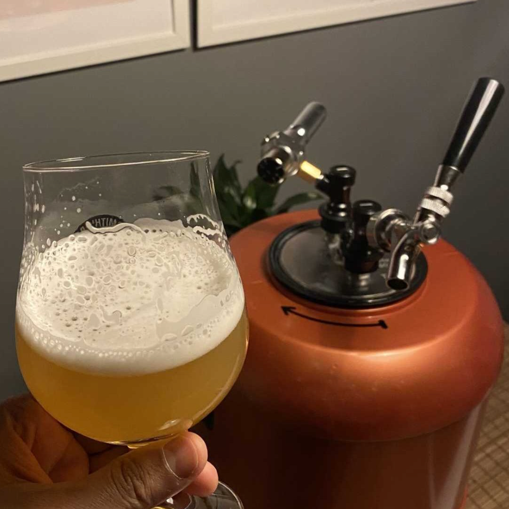 Person holding a glass of beer next to a homebrewing keg with a tap.